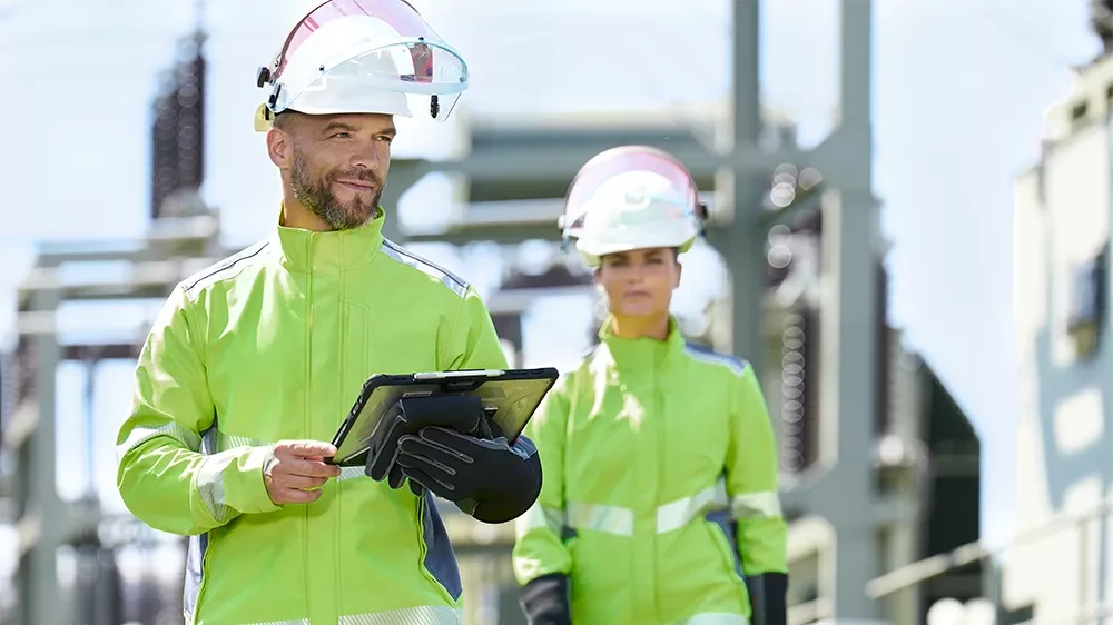 Une femme et un homme en tenue complète, composée d’un casque, de gants de protection et d’une combinaison de protection, se tiennent devant un poste de transformation.