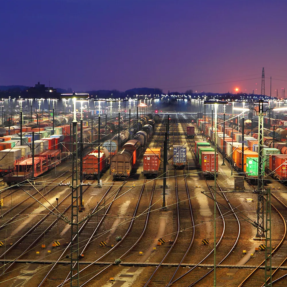 De nombreux trains de marchandises et conteneurs sont alignés dans une gare de triage très éclairée au crépuscule, avec des voies ferrées convergeant au loin et le soleil se couchant à l'horizon.