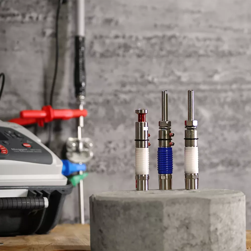 Trois objets cylindriques en métal, avec des composants rouges, bleus et blancs, sur une surface en béton devant un appareil équipé d’un panneau de commande.