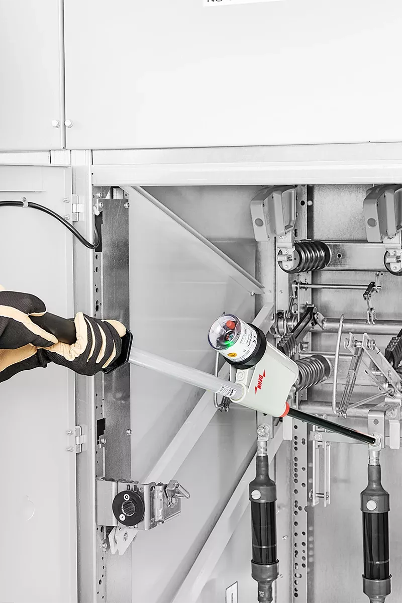Un homme en tenue de protection tient un détecteur de tension à deux mains et vérifie des connexions haute tension.