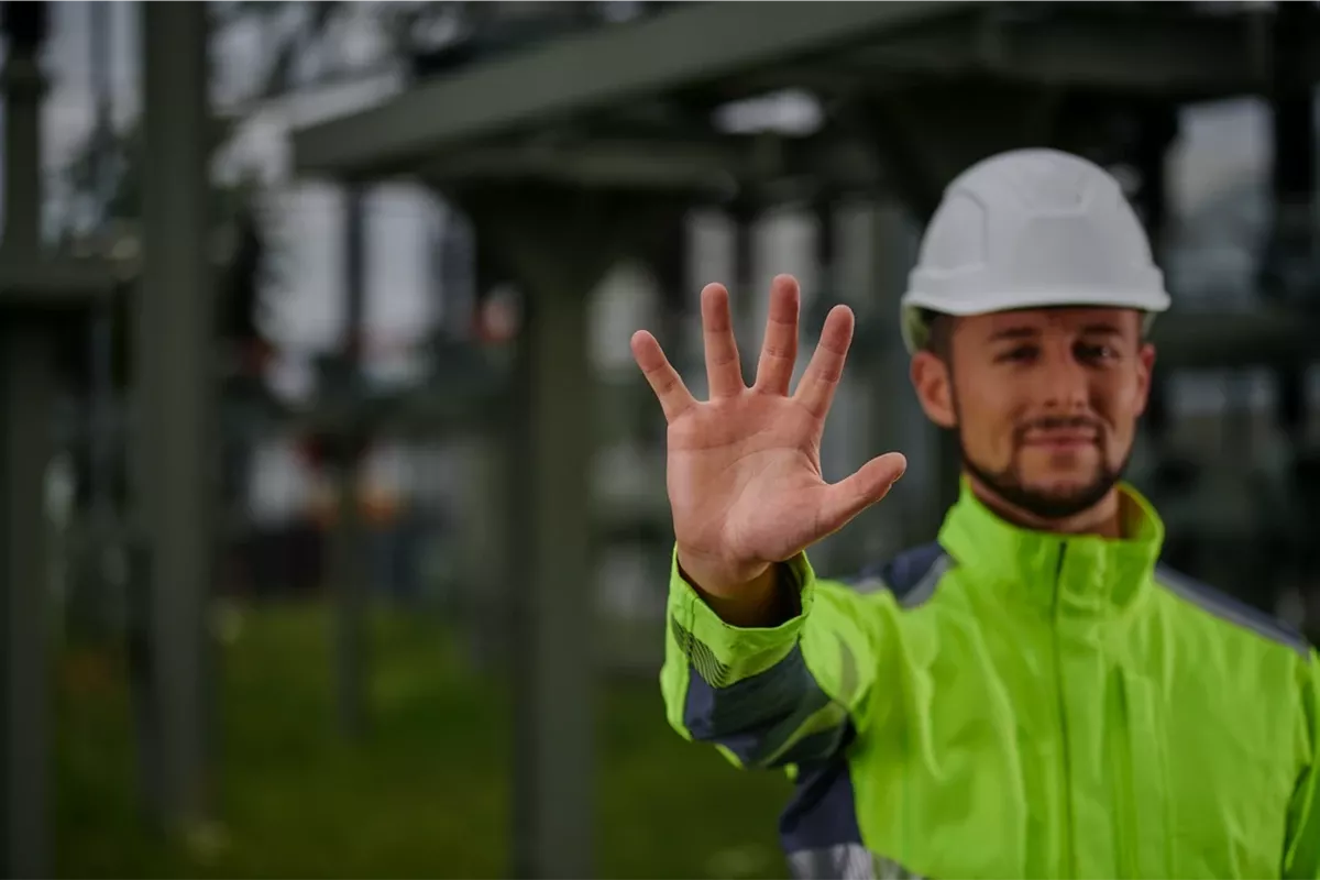 Un homme en combinaison de protection jaune et casque de protection blanc montre cinq doigts.